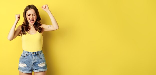 Mujer joven entusiasta gritando y animando regocijándose levantando las manos y gritando de alegría cantando de pie sobre fondo amarillo