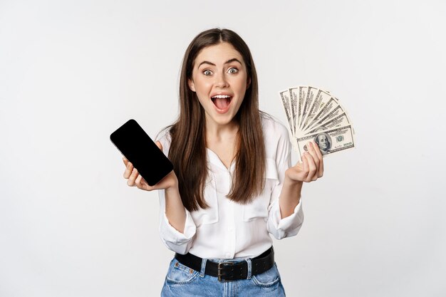 Mujer joven entusiasta que gana dinero, muestra la interfaz de la aplicación de teléfono inteligente y dinero en efectivo, microcrédito, concepto de premio, de pie sobre fondo blanco.