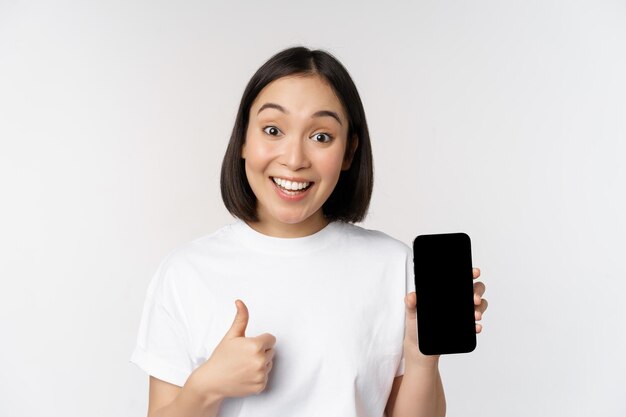 Mujer joven entusiasta que muestra el pulgar hacia arriba y la pantalla del teléfono móvil de pie en camiseta sobre fondo blanco Copiar espacio