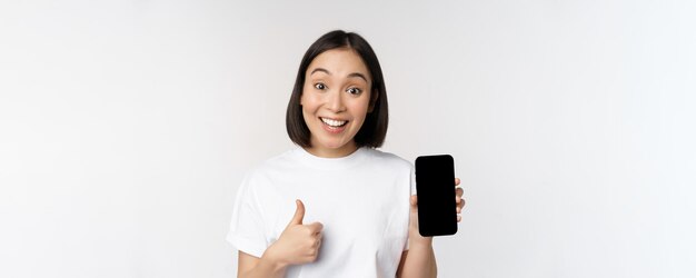 Mujer joven entusiasta que muestra el pulgar hacia arriba y la pantalla del teléfono móvil de pie en camiseta sobre fondo blanco.