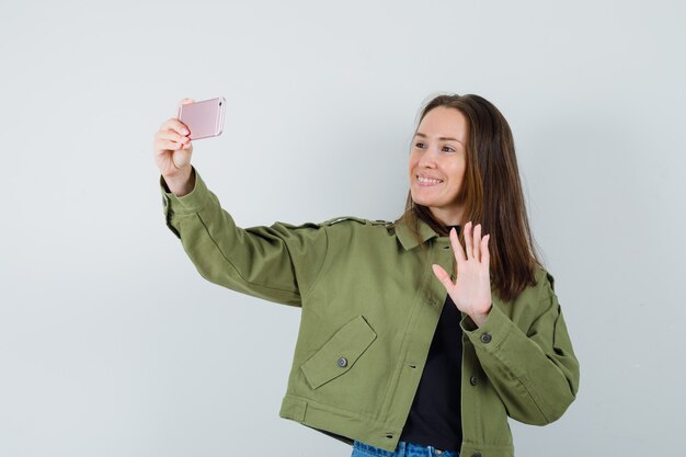 Foto gratuita mujer joven enviando saludos a alguien por teléfono con chaqueta verde y mirando contento, vista frontal.