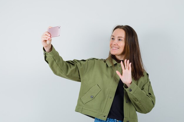 Mujer joven enviando saludos a alguien por teléfono con chaqueta verde y mirando contento, vista frontal.