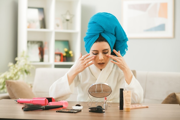 Mujer joven envuelto el cabello en una toalla aplicando crema tonificante sentado a la mesa con herramientas de maquillaje en la sala de estar