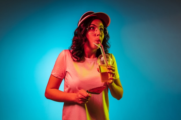 Mujer joven escalofriante con una gorra sosteniendo dulces y bebida sobre estudio de neón azul de moda