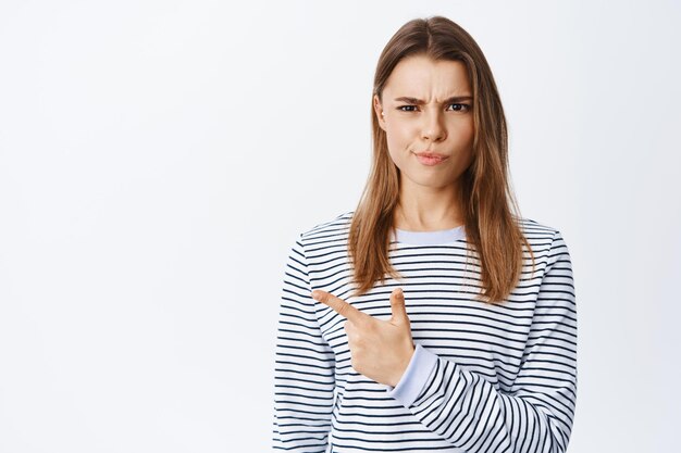 Mujer joven escéptica y sospechosa con el ceño fruncido y una sonrisa burlona, señalando con el dedo a la izquierda en la pancarta del logotipo, quejándose de algo malo, blanco