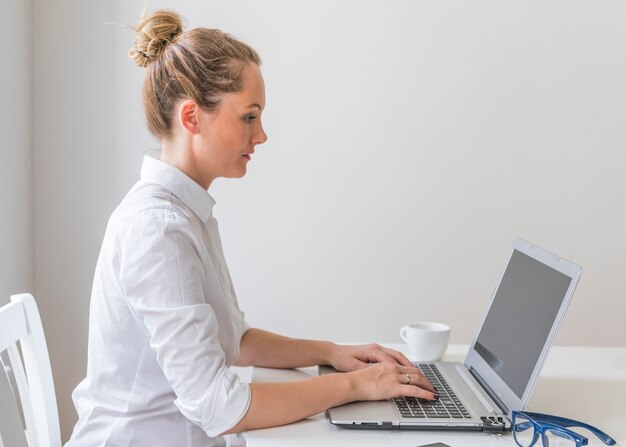 Mujer joven escribiendo en la computadora portátil con taza y anteojos en la mesa