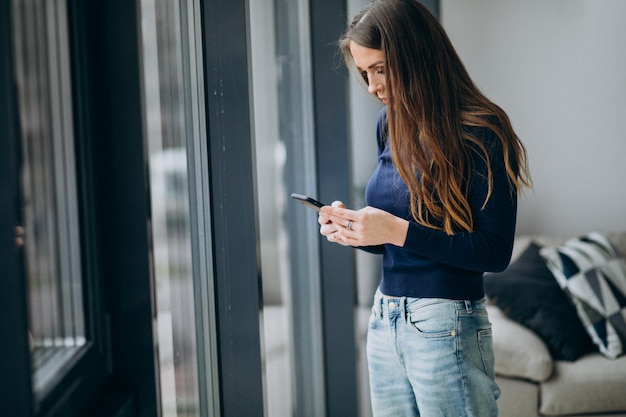 Foto gratuita mujer joven escribiendo el teclado del teléfono junto a la ventana