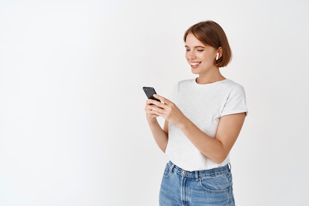 Mujer joven escucha música en auriculares inalámbricos, mirando el mensaje en el teléfono celular, leyendo la pantalla y sonriendo, de pie contra la pared blanca
