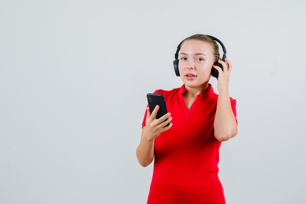 Mujer joven escuchando con auriculares en el teléfono móvil en camiseta roja y mirando positivo