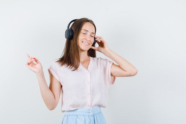 Mujer joven escuchando música con auriculares en camiseta, falda y mirando relajado, vista frontal.