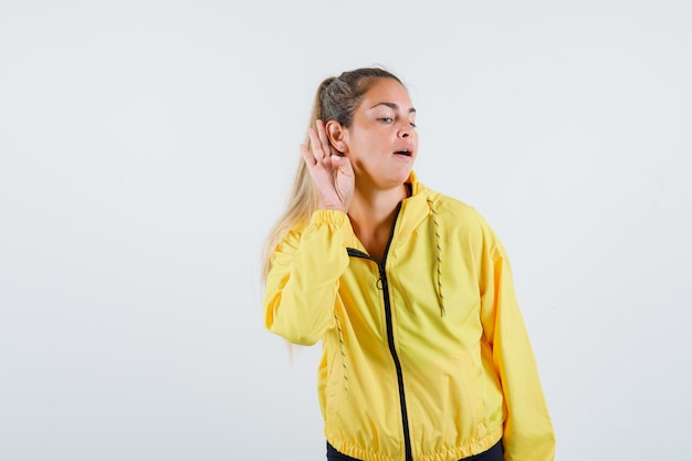 Mujer joven escuchando voces en impermeable amarillo y mirando con cuidado