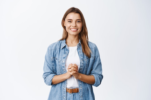 Mujer joven esperanzada con cabello rubio, tomados de la mano juntos y sonriendo cortésmente, esperando la oportunidad, lista para ayudar al cliente, de pie contra la pared blanca