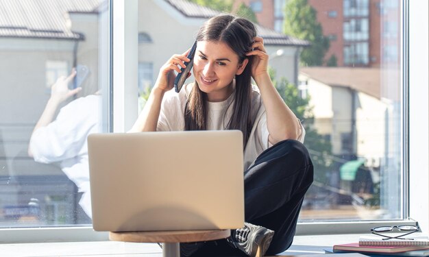 Una mujer joven está hablando en un teléfono inteligente y mirando la pantalla de una computadora portátil