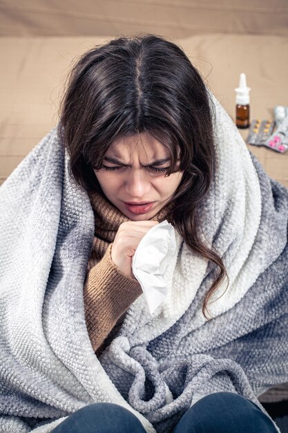 Una mujer joven está tosiendo mientras está sentada en su casa envuelta en una manta