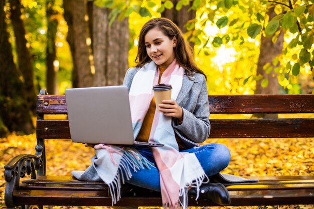 La mujer joven está usando la computadora portátil en un parque en un día del otoño.