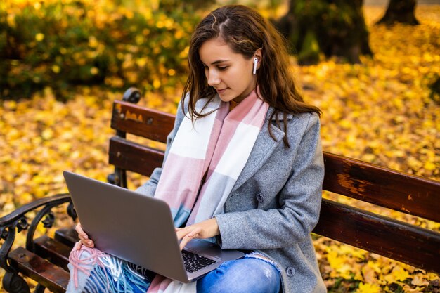 La mujer joven está usando la computadora portátil en un parque en un día del otoño.