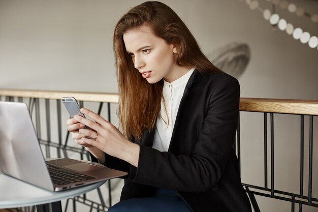 Mujer joven con estilo en la cafetería, tomando fotografías de la pantalla del portátil mediante teléfono móvil
