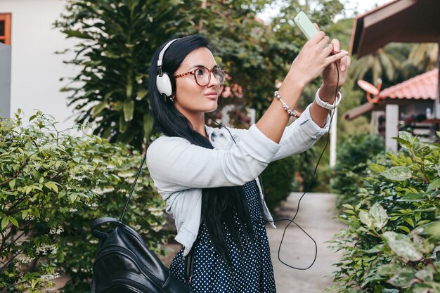 Mujer joven con estilo caminando con teléfono inteligente, escuchando música en auriculares, tomando fotos, estilo denim vintage, vacaciones de verano