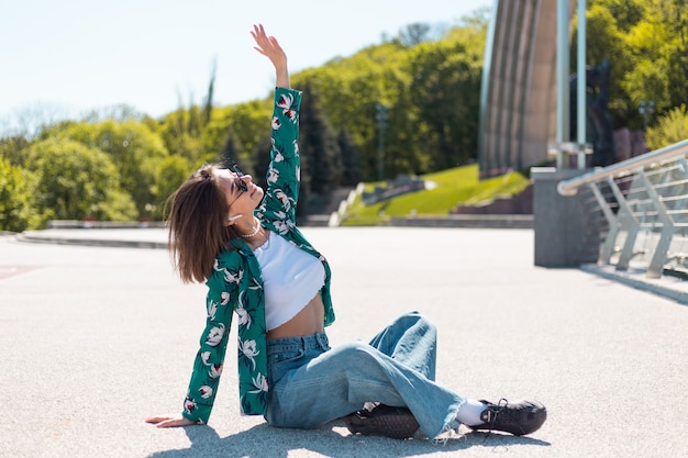 Mujer joven con estilo en camisa verde casual en día soleado posando se sienta en el puente auriculares inalámbricos bluetooth en los oídos