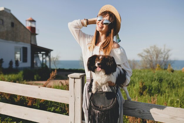 Mujer joven con estilo en el campo, sosteniendo un perro, feliz estado de ánimo positivo, verano, sombrero de paja, traje de estilo bohemio, gafas de sol, sonriente, feliz, soleado