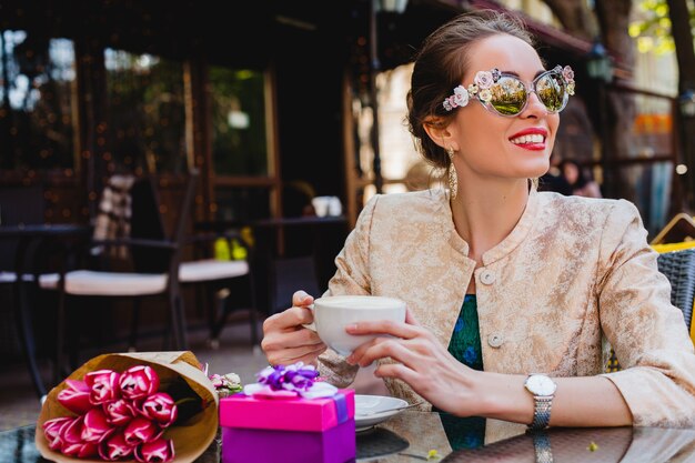 Mujer joven con estilo, gafas de sol de moda sentado en la cafetería, sosteniendo una taza de capuchino, sonriendo