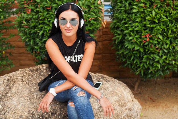 Mujer joven con estilo hipster en camiseta negra, jeans, escuchando música en auriculares, divirtiéndose, posando, divertido, sonriendo