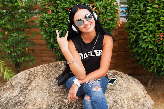 Mujer joven con estilo hipster en camiseta negra, jeans, escuchando música en auriculares, divirtiéndose, posando, divertido, sonriendo