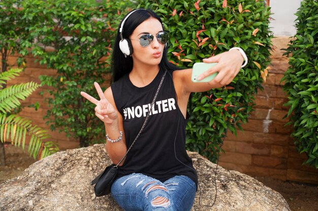 Mujer joven con estilo hipster en camiseta negra, jeans, escuchando música en auriculares, divirtiéndose, posando, tomando fotos selfie en el teléfono, mostrando el signo de la paz, expresión de la cara divertida