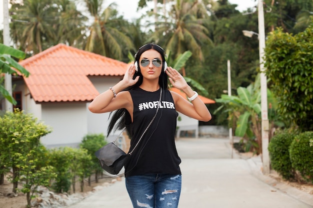 Mujer joven con estilo hipster en camiseta negra, jeans, escuchar música en auriculares, divertirse, caminar en la calle, vacaciones de verano, disfrutar