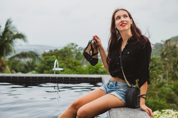 Mujer joven con estilo hipster, sentada en la piscina, vacaciones tropicales, despreocupada, belleza natural, con zapatos en sus manos, labios rojos