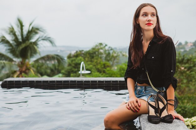 Mujer joven con estilo hipster, sentada en la piscina, vacaciones tropicales, despreocupada, belleza natural, con zapatos en sus manos, labios rojos