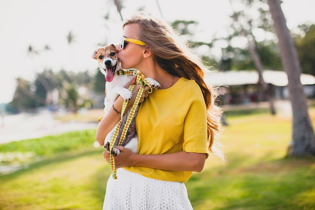 Mujer joven con estilo hipster sosteniendo caminar jugando perro cachorro jack russell, parque tropical, sonriendo y divirtiéndose, vacaciones, gafas de sol, gorra, camisa amarilla, arena de playa