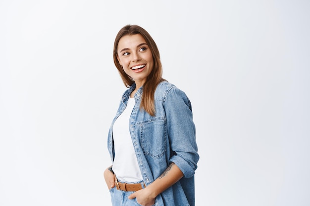 Mujer joven con estilo en ropa casual, de pie de perfil y girar la cabeza a la derecha en el espacio de la copia con publicidad, leyendo ofertas promocionales, de pie en blanco