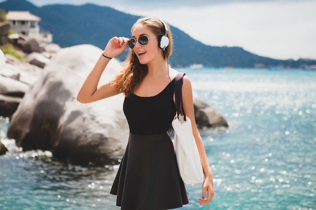 Mujer joven con estilo sexy hipster con una bolsa de compras durante las vacaciones, gafas de sol de aviador, auriculares, escuchando música, feliz, disfrutando del sol, paisaje de la laguna azul de la isla tropical