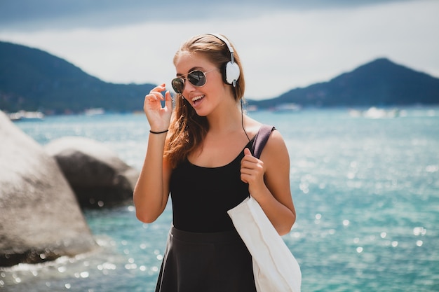 Mujer joven con estilo sexy hipster con una bolsa de compras durante las vacaciones, gafas de sol de aviador, auriculares, escuchando música, feliz, disfrutando del sol, paisaje de la laguna azul de la isla tropical