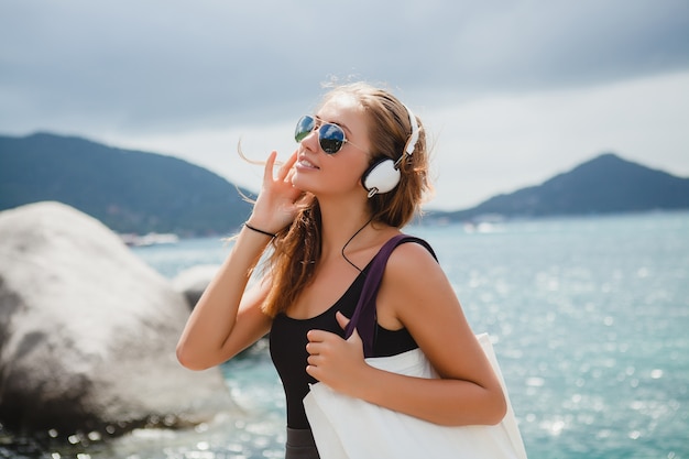 Mujer joven con estilo sexy hipster con una bolsa de compras durante las vacaciones, gafas de sol de aviador, auriculares, escuchando música, feliz, disfrutando del sol, paisaje de la laguna azul de la isla tropical