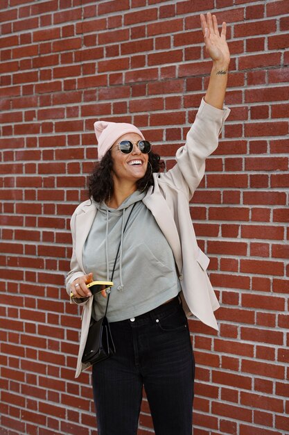 Mujer joven con estilo con smartphone contra la pared de ladrillo en la ciudad