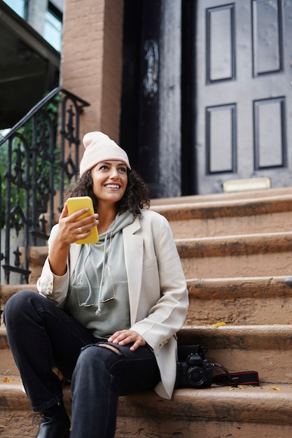 Mujer joven con estilo con smartphone mientras está sentado en las estepas de la ciudad