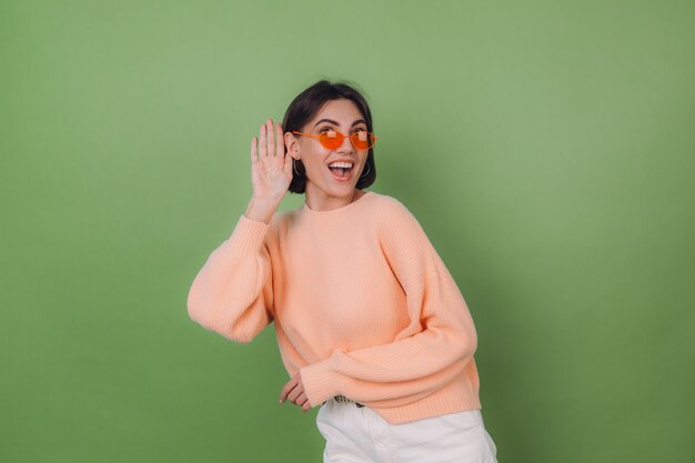 Mujer joven con estilo en suéter de melocotón casual y gafas naranjas aisladas en la pared verde oliva curioso, intenta escuchar lo que dices con la mano en el espacio de copia de oído