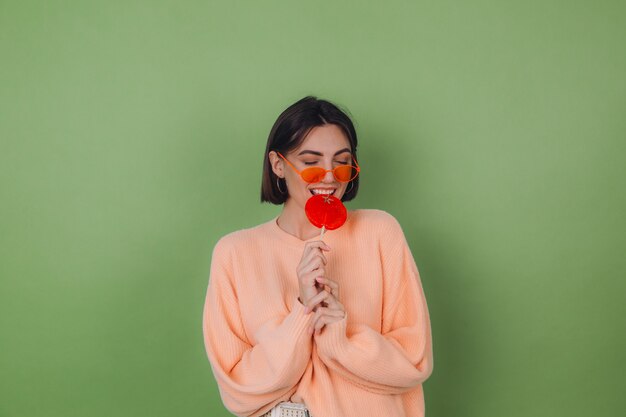 Mujer joven con estilo en suéter de melocotón casual y gafas naranjas aisladas en la pared verde oliva con espacio de copia de sonrisa positiva de piruleta naranja