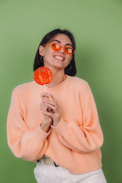 Mujer joven con estilo en suéter de melocotón casual y gafas naranjas aisladas en la pared verde oliva con espacio de copia de sonrisa positiva de piruleta naranja