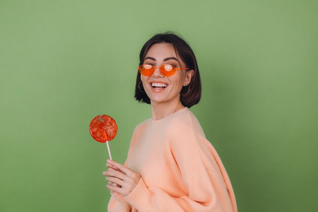 Mujer joven con estilo en suéter de melocotón casual y gafas naranjas aisladas en la pared verde oliva con espacio de copia de sonrisa positiva de piruleta naranja