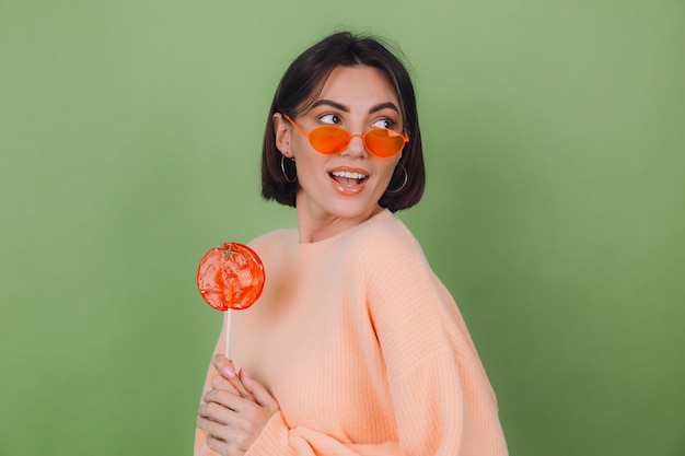 Mujer joven con estilo en suéter de melocotón casual y gafas naranjas aisladas en la pared verde oliva con espacio de copia de sonrisa positiva de piruleta naranja