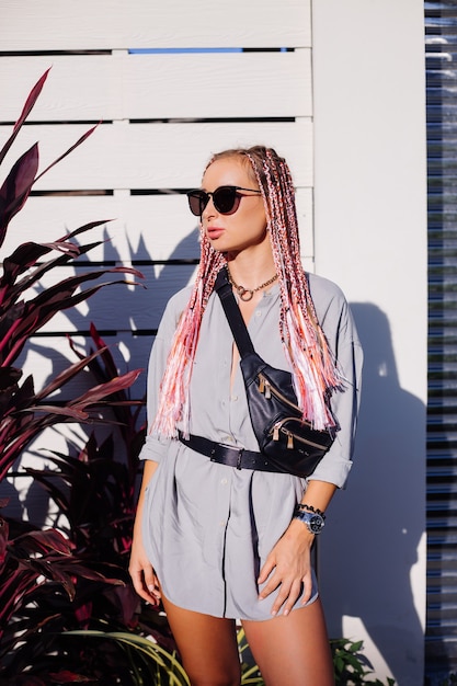 Mujer joven con estilo con trenzas rosa púrpura y riñonera negra posando al aire libre