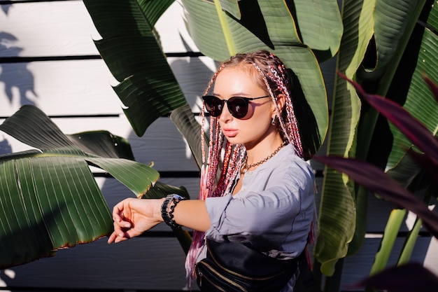 Mujer joven con estilo con trenzas rosa púrpura y riñonera negra posando al aire libre