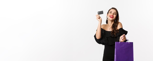 Foto gratuita mujer joven con estilo en vestido negro ir de compras con bolsa y tarjeta de crédito sonriendo complacido standi