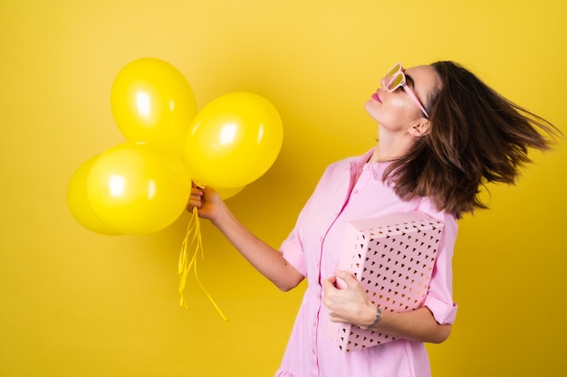 Mujer joven con estilo en un vestido rosa y gafas con una caja de regalo y globos amarillos en sus manos