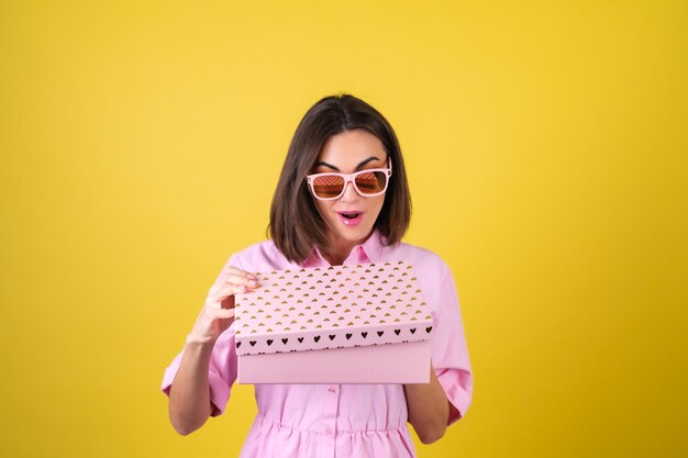 Mujer joven con estilo en un vestido rosa y gafas con una caja de regalo en sus manos