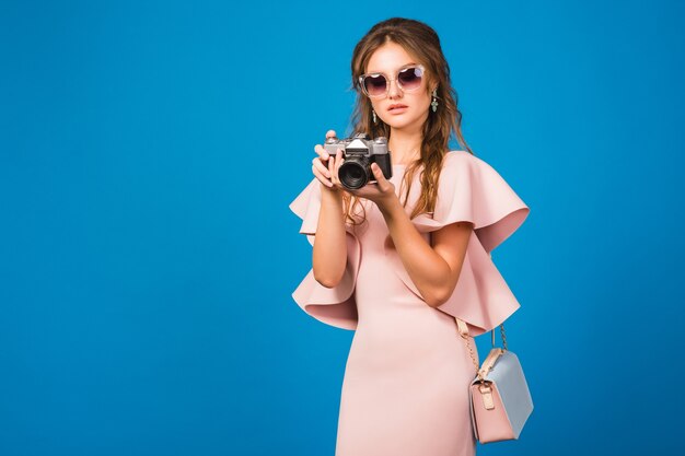 Mujer joven con estilo en vestido rosa de lujo tomando fotografías en cámara vintage