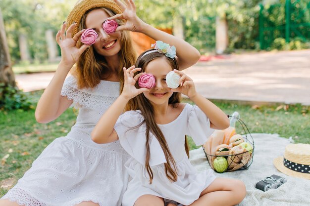 Una mujer joven con estilo vino con una hija bonita al parque para pasar el fin de semana juntos. Retrato al aire libre de niña de pelo castaño bromeando con la madre mientras come galletas en una manta.